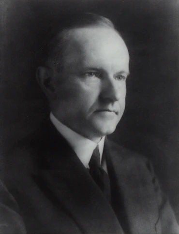 Professional portrait of a serious, confident man in formal business attire, standing with arms crossed in an office environment.