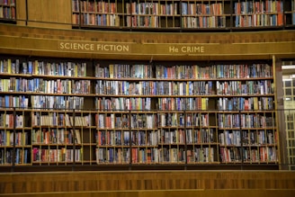 A large wooden bookshelf filled with an extensive collection of books organized into sections labeled as 'Science Fiction' and 'Crime'. The shelves are packed with colorful book spines, featuring diverse titles and authors.