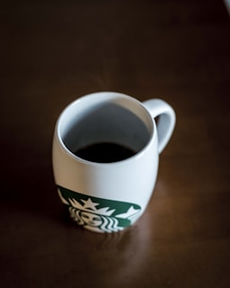 A white mug filled with black coffee is placed on a brown table. The mug features a green logo with a siren design.