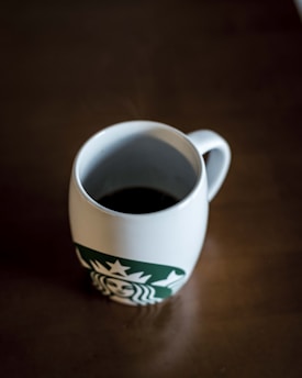 A white mug filled with black coffee is placed on a brown table. The mug features a green logo with a siren design.