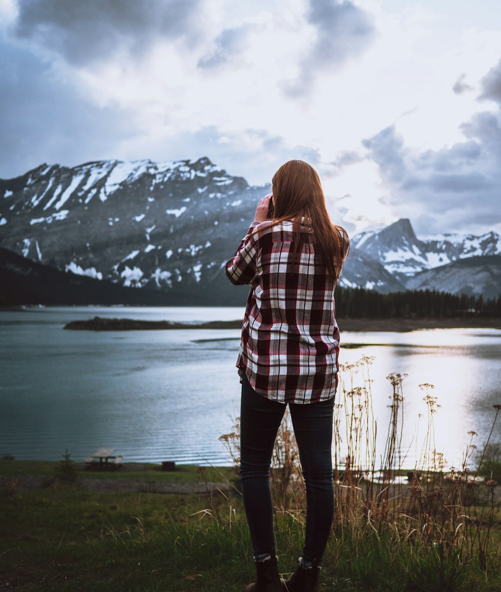 woman in plaid shirt standing on green grass near lake and snow covered mountain during daytime
