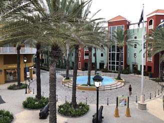 A plaza surrounded by colorful buildings with red roofs and palm trees. A circular fountain is in the center of the plaza. There are shops with visible signs, street lamps, and a woman walking nearby.