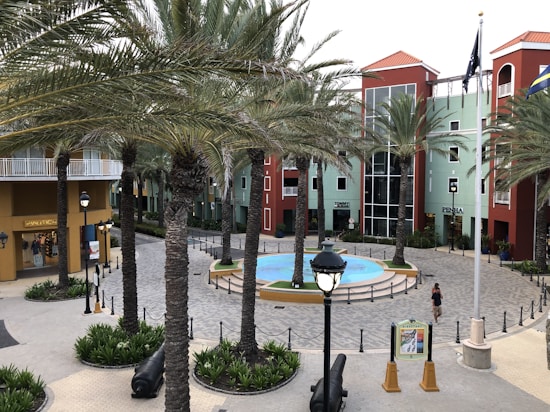 A plaza surrounded by colorful buildings with red roofs and palm trees. A circular fountain is in the center of the plaza. There are shops with visible signs, street lamps, and a woman walking nearby.