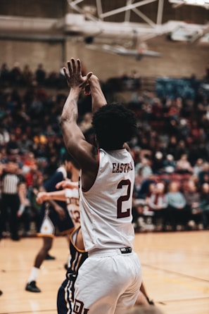 A basketball player wearing a white jersey with the number 2 is captured in mid-action, making a shot. The player has a focused posture with arms extended. In the background, there is an indoor basketball court with spectators watching the game.