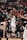 A basketball player in white uniform is actively shooting the ball in a crowded indoor arena. Spectators fill the bleachers, watching intently. The scene is dynamic, with players from the opposing team visible in the foreground.