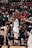 A basketball player in white uniform is actively shooting the ball in a crowded indoor arena. Spectators fill the bleachers, watching intently. The scene is dynamic, with players from the opposing team visible in the foreground.