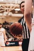 A basketball referee wearing a black and white striped shirt holds a basketball with both hands, standing between two players on a court. In the background, blurred spectators and a scoreboard are visible.