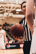 A basketball referee wearing a black and white striped shirt holds a basketball with both hands, standing between two players on a court. In the background, blurred spectators and a scoreboard are visible.