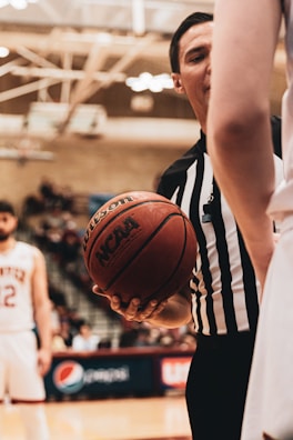 A basketball referee wearing a black and white striped shirt holds a basketball with both hands, standing between two players on a court. In the background, blurred spectators and a scoreboard are visible.