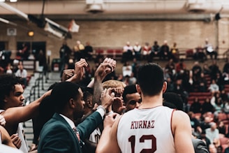 A group of basketball players huddle together in a gymnasium, raising their hands in unison with a sense of unity and teamwork. They appear to be preparing or celebrating during an intense game, with a crowd of spectators in the background watching from the bleachers.