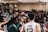 A group of basketball players huddle together in a gymnasium, raising their hands in unison with a sense of unity and teamwork. They appear to be preparing or celebrating during an intense game, with a crowd of spectators in the background watching from the bleachers.