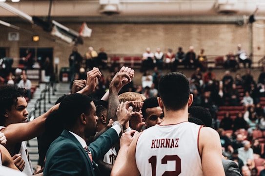 A group of basketball players huddle together in a gymnasium, raising their hands in unison with a sense of unity and teamwork. They appear to be preparing or celebrating during an intense game, with a crowd of spectators in the background watching from the bleachers.