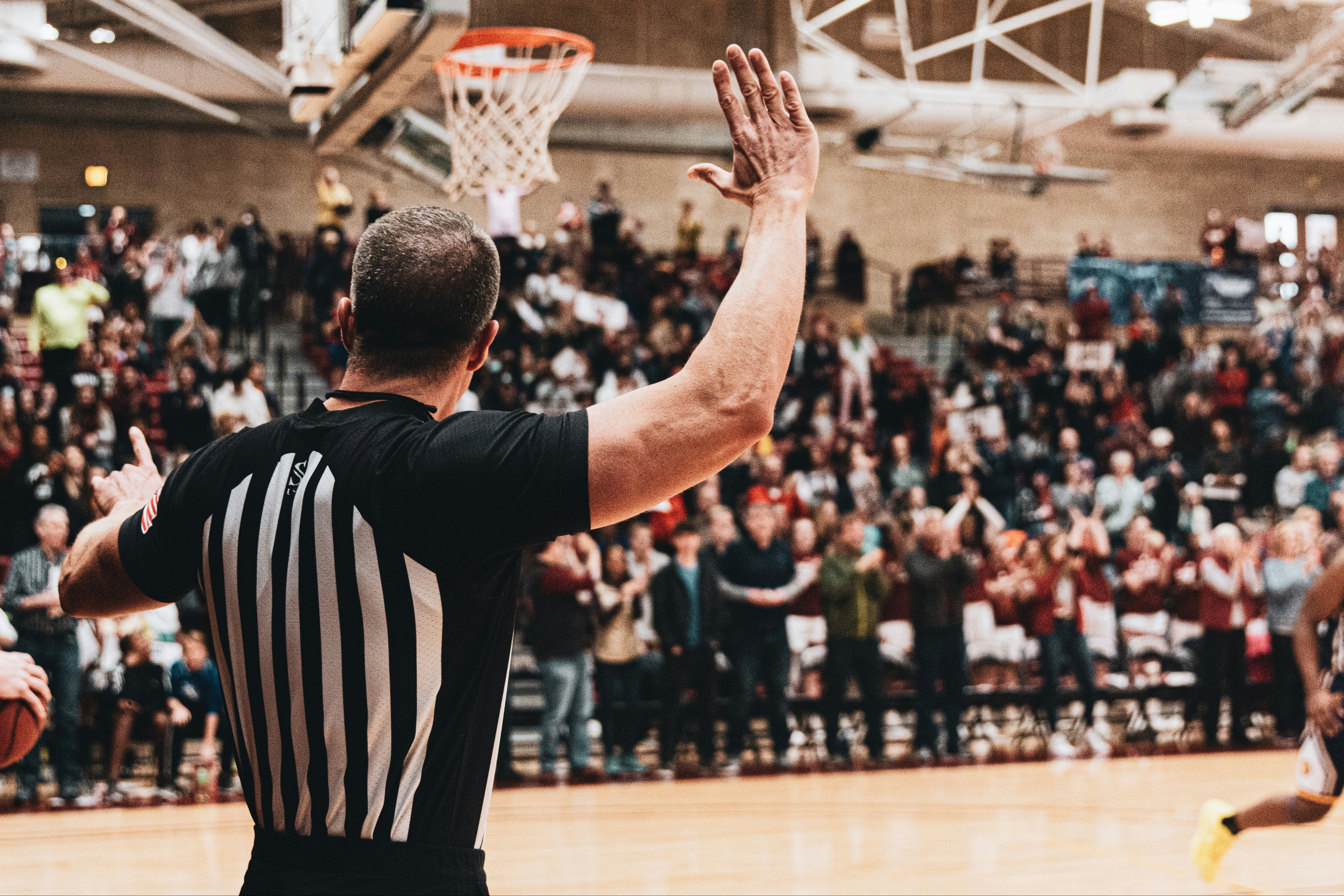 A basketball referee gestures during a game with a cheering crowd in the background.