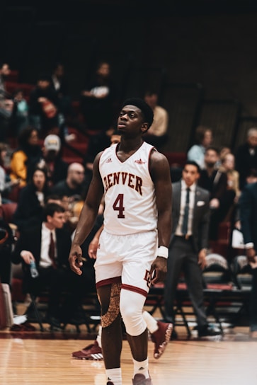 A basketball player wearing a white jersey with the number 4 and the name 'Denver' is on a basketball court. He has a knee brace on his left knee and appears to be walking. The background shows a blurred crowd sitting and standing, suggesting a game or match setting.
