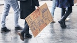 People walking on a wet pavement, carrying a protest sign demanding equal pay and bigger pockets, highlighting gender equality issues.