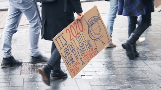 People walking on a wet pavement, carrying a protest sign demanding equal pay and bigger pockets, highlighting gender equality issues.