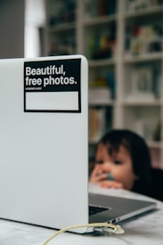A laptop with a sticker saying 'Beautiful, free photos' is in the foreground, partially blocking a blurred background. A young child holding something in their mouth is visible behind the laptop, adding a playful element to the image. The setting appears to be domestic, with shelves filled with colorful books in the background.