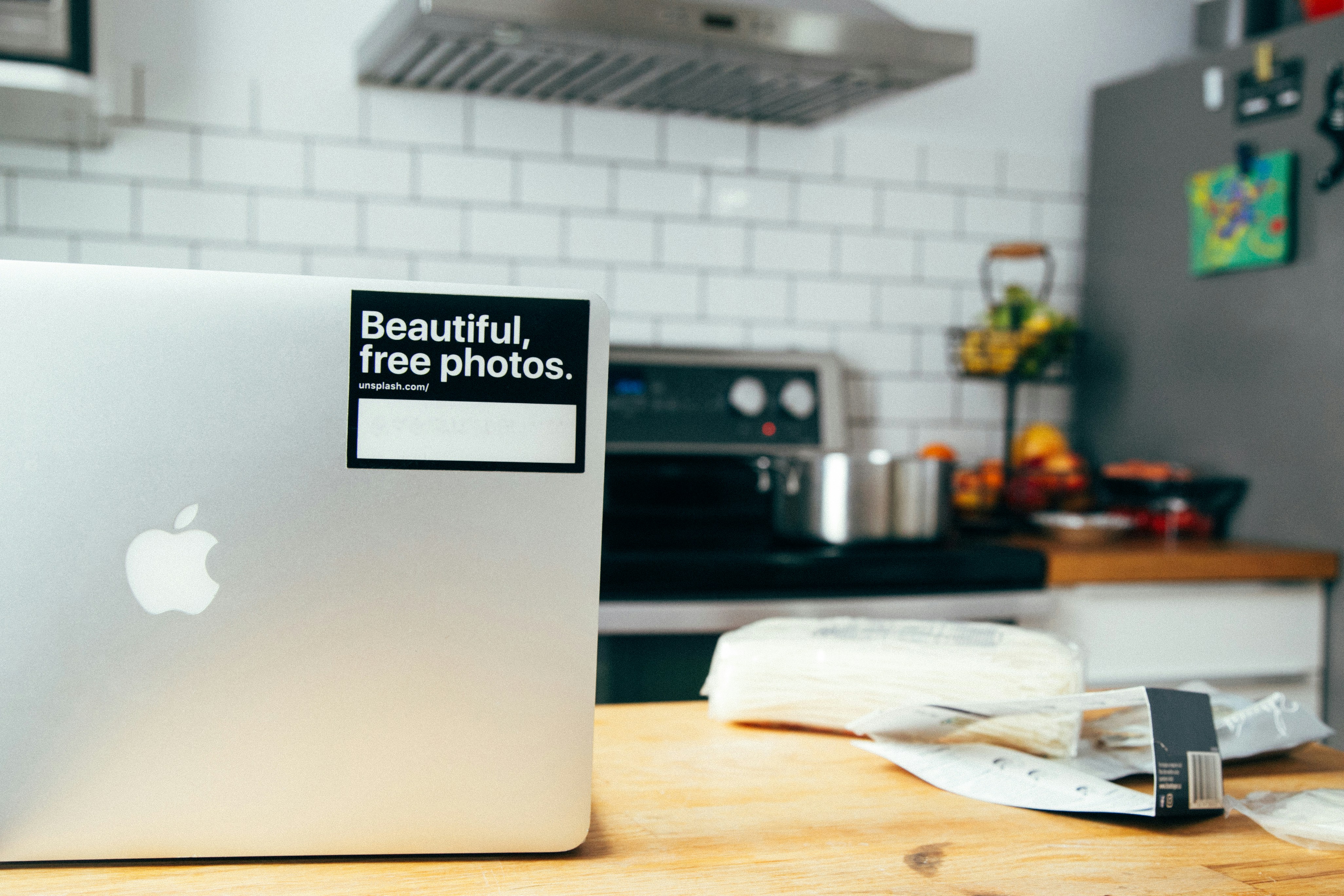 A modern kitchen setting with a silver laptop on a wooden table, featuring a sticker that reads 'Beautiful, free photos.' The background includes a stove with a stainless steel pot, a fruit bowl with various fruits, a tiled backsplash, and a refrigerator with magnets.
