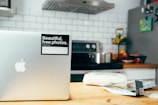 A modern kitchen setting with a silver laptop on a wooden table, featuring a sticker that reads 'Beautiful, free photos.' The background includes a stove with a stainless steel pot, a fruit bowl with various fruits, a tiled backsplash, and a refrigerator with magnets.