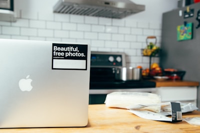 A modern kitchen setting with a silver laptop on a wooden table, featuring a sticker that reads 'Beautiful, free photos.' The background includes a stove with a stainless steel pot, a fruit bowl with various fruits, a tiled backsplash, and a refrigerator with magnets.
