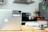 A modern kitchen setting with a silver laptop on a wooden table, featuring a sticker that reads 'Beautiful, free photos.' The background includes a stove with a stainless steel pot, a fruit bowl with various fruits, a tiled backsplash, and a refrigerator with magnets.