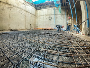 A construction site with two workers wearing red hard hats, one bending over and the other standing, surrounded by a framework of steel rebar laid on the ground within a concrete structure. The site is enclosed by tall concrete walls, and blue and green fabric sheets are partially covering the top.