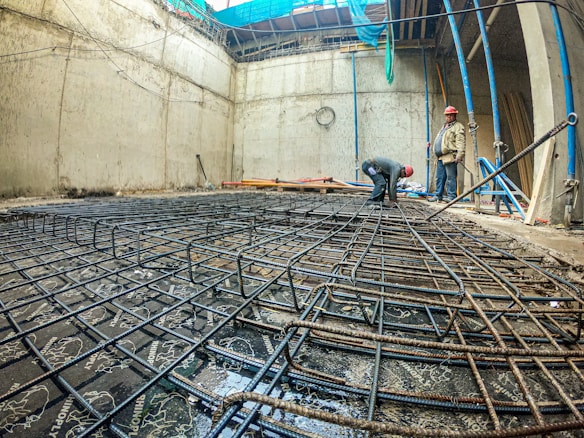 A construction site with two workers wearing red hard hats, one bending over and the other standing, surrounded by a framework of steel rebar laid on the ground within a concrete structure. The site is enclosed by tall concrete walls, and blue and green fabric sheets are partially covering the top.