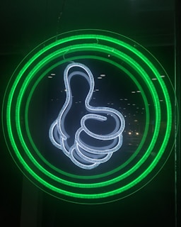 A neon light display features a thumbs-up symbol outlined in bright white. Surrounding the hand gesture are concentric circles glowing in green, set against a dark background.
