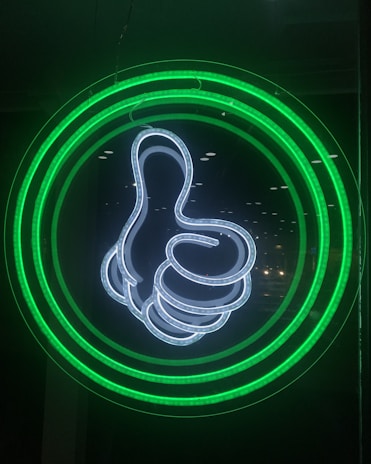 A neon light display features a thumbs-up symbol outlined in bright white. Surrounding the hand gesture are concentric circles glowing in green, set against a dark background.