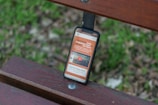 A smartphone rests on a wooden bench with a grassy background. The phone screen displays a website with orange and white text and includes a video thumbnail.