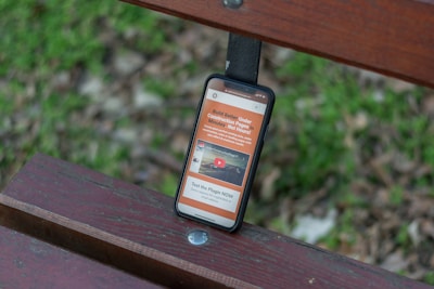 A smartphone rests on a wooden bench with a grassy background. The phone screen displays a website with orange and white text and includes a video thumbnail.