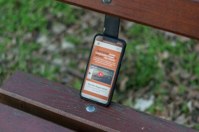 A smartphone rests on a wooden bench with a grassy background. The phone screen displays a website with orange and white text and includes a video thumbnail.