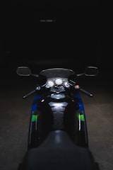 A motorcycle is parked in a dimly lit environment, featuring a close-up view of the handlebars, mirrors, and dashboard. The black seat and parts are contrasted by blue and green elements on the body of the bike.
