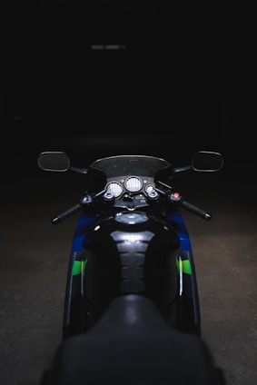 A motorcycle is parked in a dimly lit environment, featuring a close-up view of the handlebars, mirrors, and dashboard. The black seat and parts are contrasted by blue and green elements on the body of the bike.
