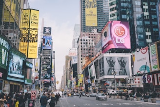 A bustling urban scene filled with numerous bright and colorful billboards and advertisements. Large signage for brands and Broadway shows are prominently displayed on buildings lining the busy street. People are walking and cars are passing through the intersection, capturing the vibrant energy of a city.