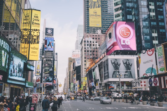 A bustling urban scene filled with numerous bright and colorful billboards and advertisements. Large signage for brands and Broadway shows are prominently displayed on buildings lining the busy street. People are walking and cars are passing through the intersection, capturing the vibrant energy of a city.