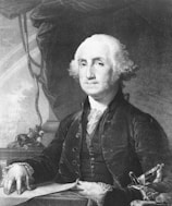 A monochrome portrait of a distinguished man dressed in 18th-century formal attire, featuring a dark jacket with buttons and lace cuffs. The subject has a solemn expression and is posed seated at a desk with one hand resting on a document. The background includes draped fabric and a table with ornate detailing.