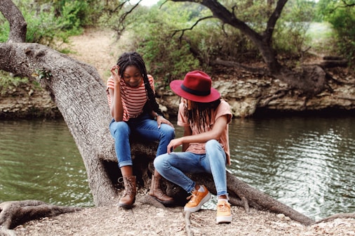 woman in red hat and blue denim jeans sitting on tree trunk beside river during daytime