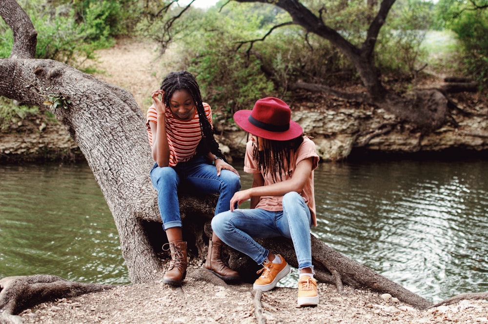 赤い帽子とブルーデニムのジーンズを履いた女性が昼間、川のそばの木の幹に座っている