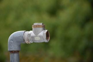 A metal pipe with a valve is positioned in the foreground against a blurred green background. The pipe appears to be weathered, with visible rust and worn threads. A piece of cloth or twine is wrapped around the joint, possibly for sealing or support.
