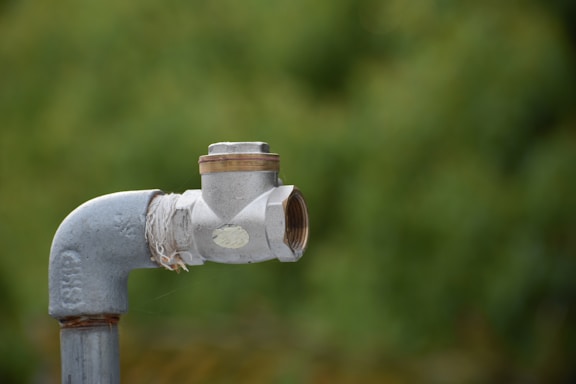 A metal pipe with a valve is positioned in the foreground against a blurred green background. The pipe appears to be weathered, with visible rust and worn threads. A piece of cloth or twine is wrapped around the joint, possibly for sealing or support.