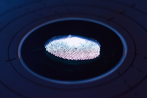 A close-up of a fingerprint illuminated by a blue and purple light, set against a dark background with faint concentric circles. The fingerprint's ridges are highlighted, giving a sense of depth and texture.
