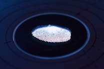 A close-up of a fingerprint illuminated by a blue and purple light, set against a dark background with faint concentric circles. The fingerprint's ridges are highlighted, giving a sense of depth and texture.