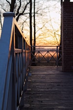 A wooden deck with a railing on the left side extends towards a sunset in the background. Bare trees without leaves are silhouetted against the orange and yellow hues of the sky. A brick column is visible on the right edge, and the deck is scattered with small debris.