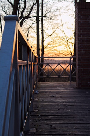 A wooden deck with a railing on the left side extends towards a sunset in the background. Bare trees without leaves are silhouetted against the orange and yellow hues of the sky. A brick column is visible on the right edge, and the deck is scattered with small debris.