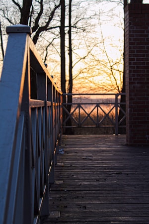 A wooden deck with a railing on the left side extends towards a sunset in the background. Bare trees without leaves are silhouetted against the orange and yellow hues of the sky. A brick column is visible on the right edge, and the deck is scattered with small debris.