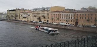 A canal in an urban setting with a tour boat cruising through the water. The backdrop consists of a row of historic buildings with classic architectural features, such as arched windows and ornate balconies. Vehicles are parked along the street adjacent to the canal. The sky is overcast, contributing to a muted atmosphere.