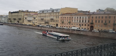 A canal in an urban setting with a tour boat cruising through the water. The backdrop consists of a row of historic buildings with classic architectural features, such as arched windows and ornate balconies. Vehicles are parked along the street adjacent to the canal. The sky is overcast, contributing to a muted atmosphere.