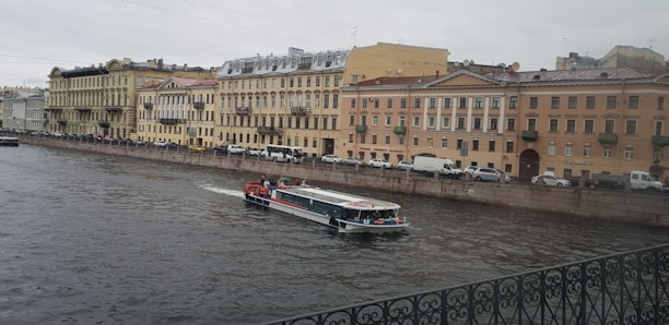 A canal in an urban setting with a tour boat cruising through the water. The backdrop consists of a row of historic buildings with classic architectural features, such as arched windows and ornate balconies. Vehicles are parked along the street adjacent to the canal. The sky is overcast, contributing to a muted atmosphere.