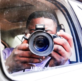 A person is holding a DSLR camera, capturing an image through the side mirror of a vehicle. The focus is on the camera lens, and the person's hands are visible. The background shows the interior of the car and a reflection.